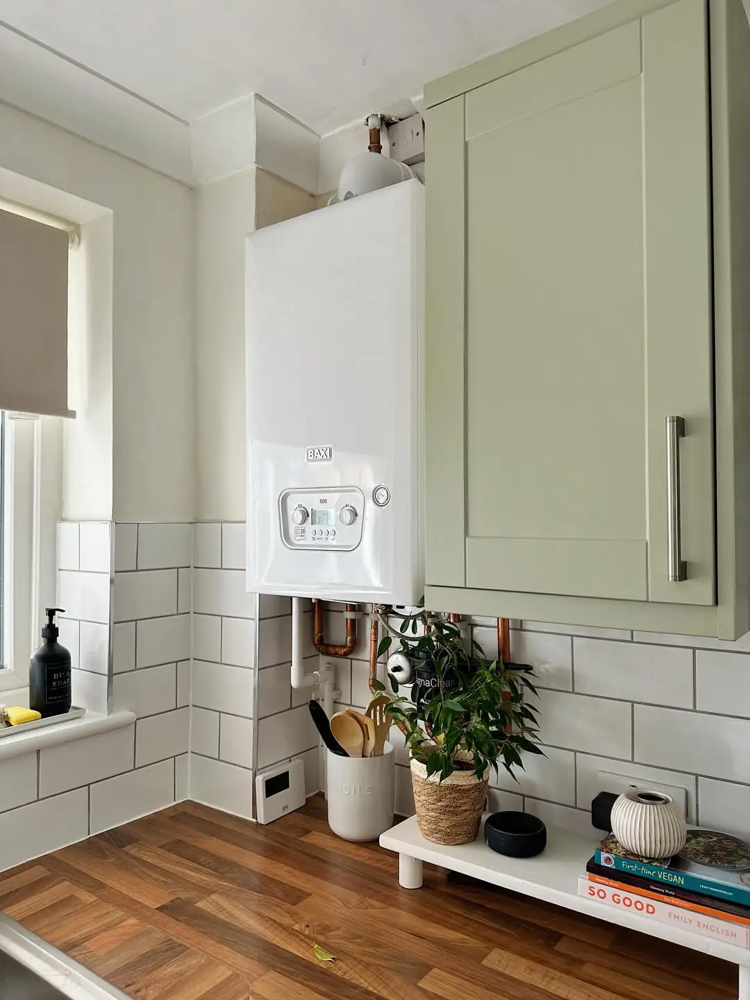 Gas boiler and central-heating pipework installation by J. Copland. Boiler is located in a kitchen in the corner next to an olive green cupboard with utensils and a house plant below it on the oak work top.